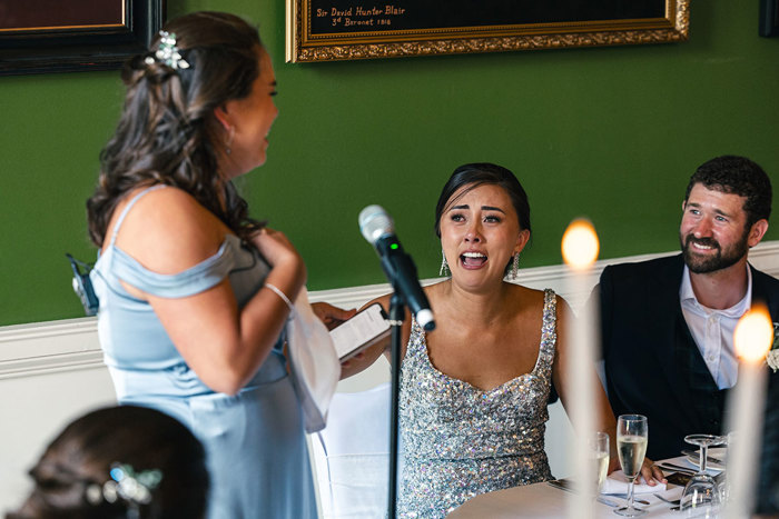 a bride seated at a table wearing a silver sparkly dress sitting next to a person wearing a dark blue suit gasps in emotion while looking at a bridesmaid who is standing next to a microphone