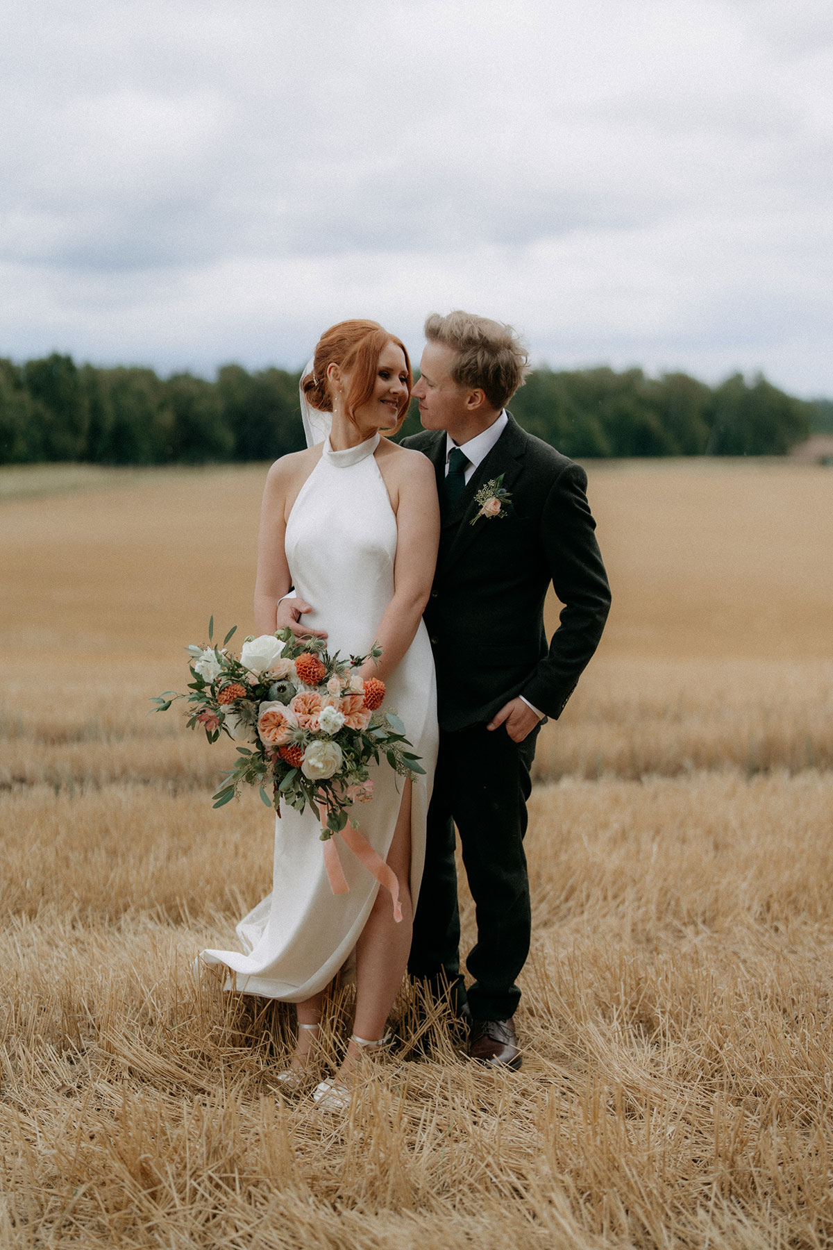 Bride and groom standing together in golden field holding peach and white wedding bouquet