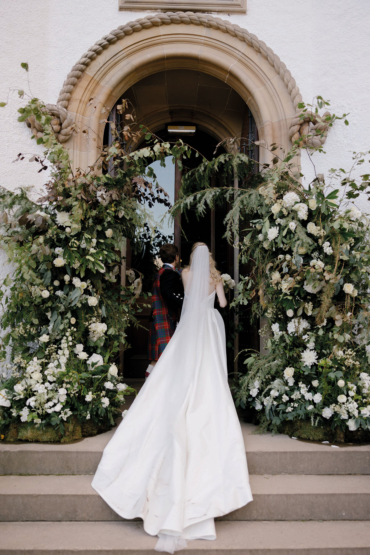 A bride in a long satin gown and veil walks with her groom through an abundant white and green floral arch at a grand stone doorway