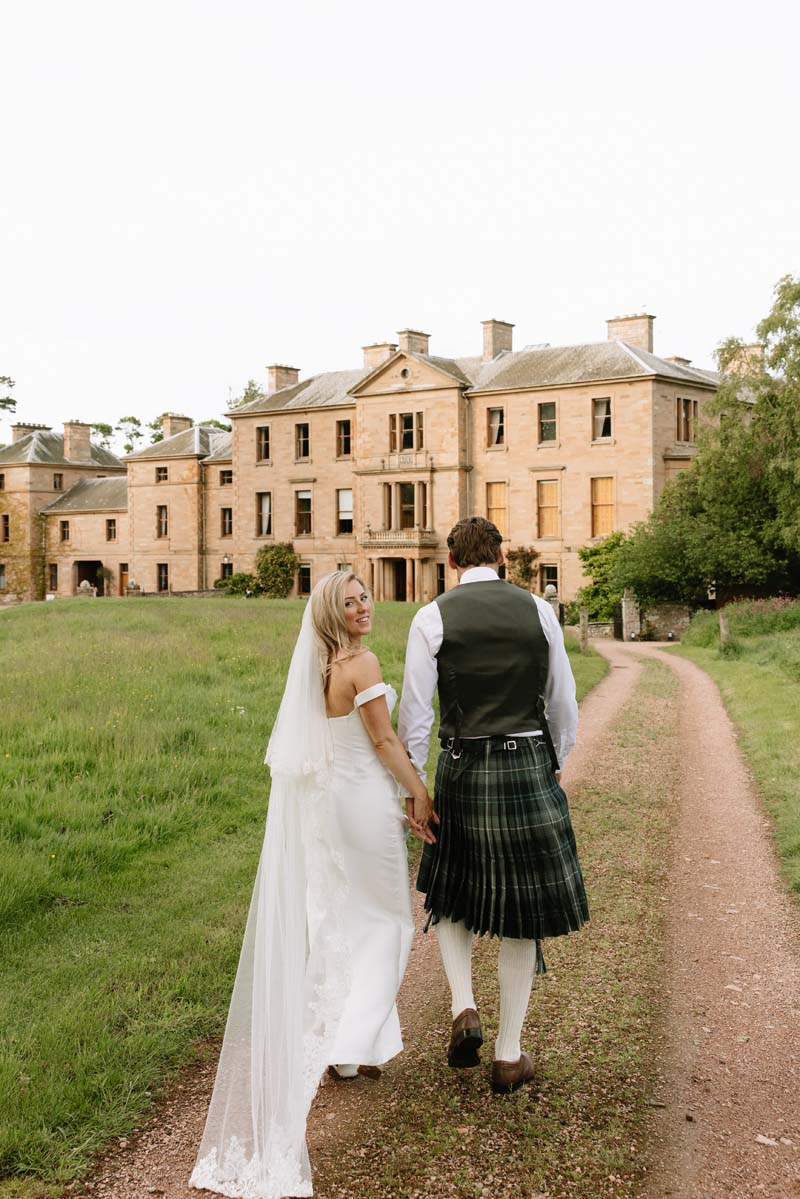 Lia and Louis walk hand in hand through the grounds with Cambo House in the background.