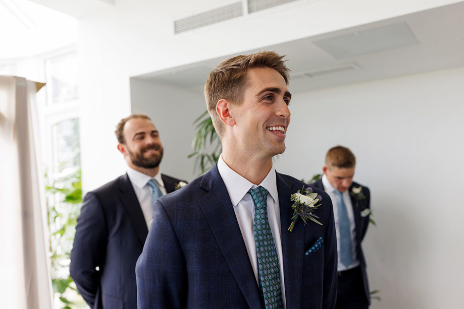 A joyous groom in a blue suit smiling brightly at his Old Course Hotel wedding, accompanied by groomsmen in the background sharing in the happy occasion, immortalized by Duke Photography