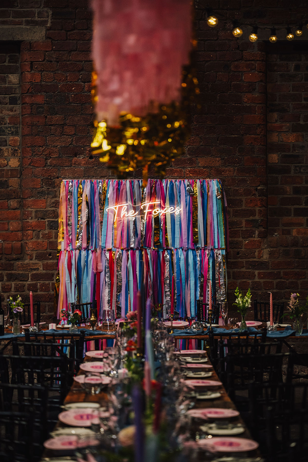 Long wooden wedding tables set with pastel plates, tapered candles and floral arrangements, with colourful pink, blue and silver streamers hanging overhead and a neon “The Foxes” sign in the background.