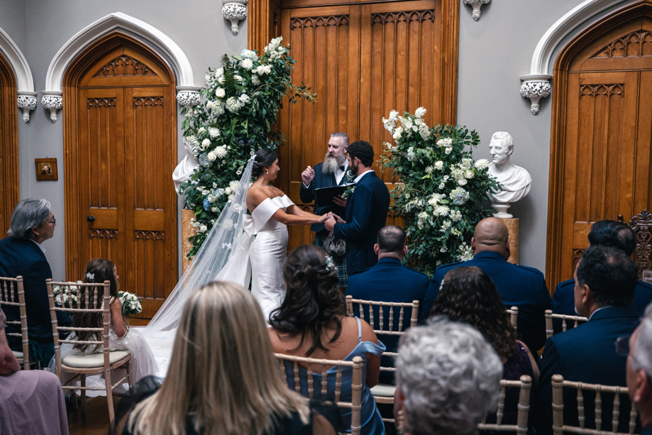 A bride and groom getting married with a celebrant standing between two flower pillars and grand wooden doors as an audience looks on