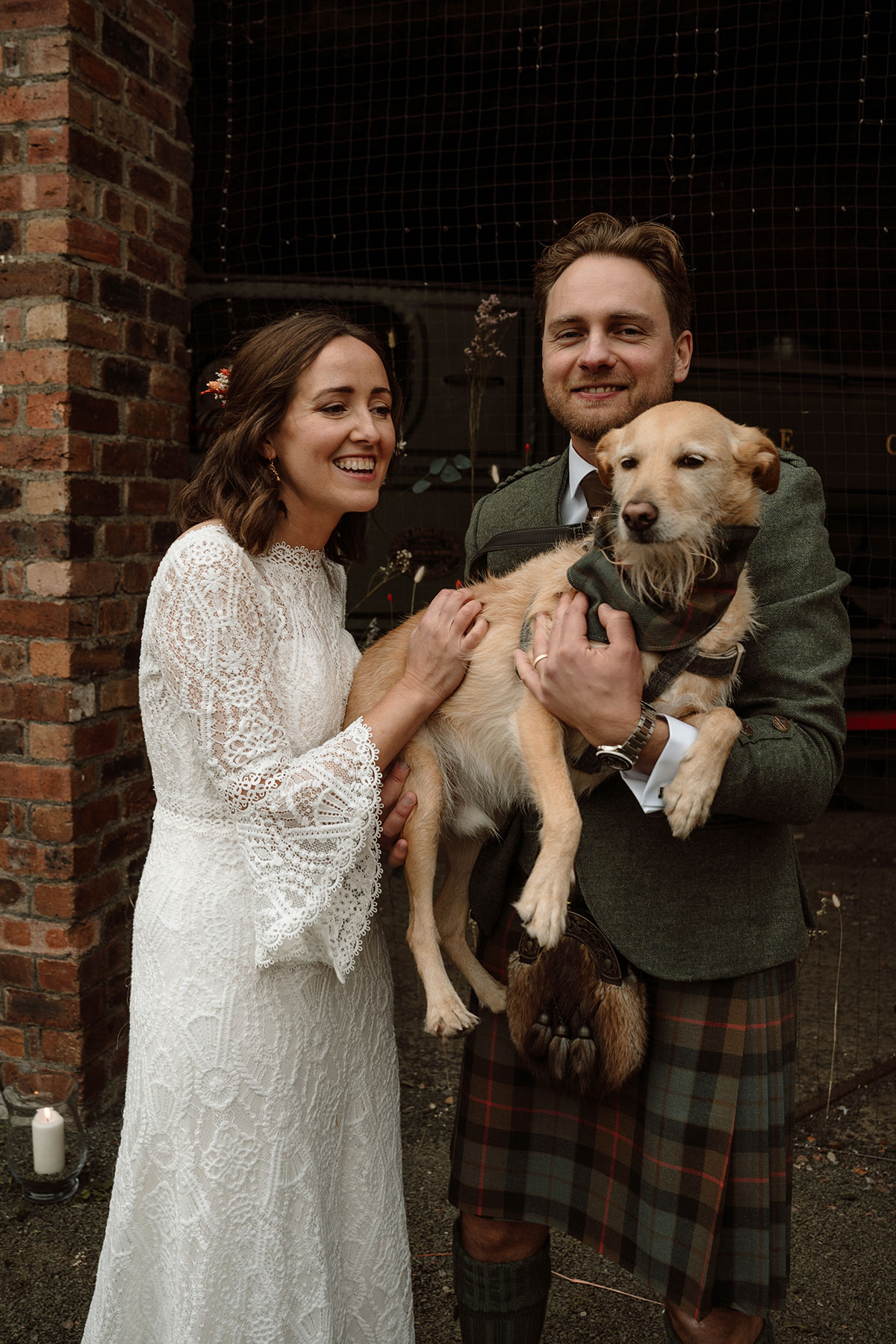 Bride and groom pose with their dog outside the wedding venue, with the groom wearing a tartan kilt and the bride in a long-sleeved lace wedding dress