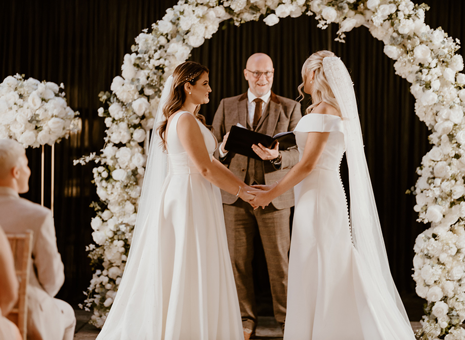 two brides stand holding hands beside a white floral arch during their wedding ceremony with celebrant standing between them reading from a binder