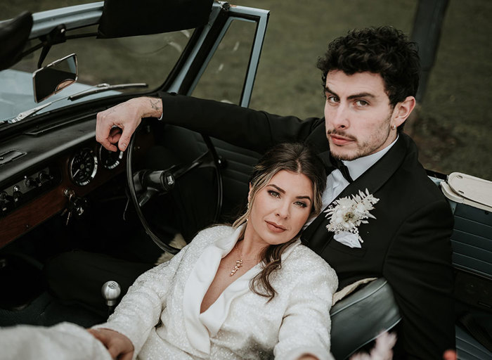 A woman in a white suit leans against a man in a black suit as they sit in convertible car