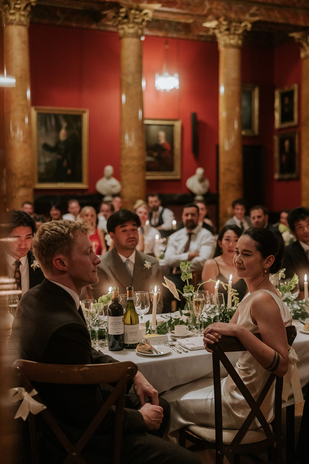 Bride and groom seated at dinner table, surrounded by guests in grand hall.