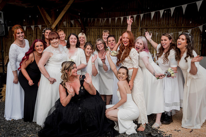 A Bride Wearing Black Crouches Down In A Wooden Building With Bunting Hanging On The Walls While Surrounded By A Group Of Women Wearing White And Ivory Dresses