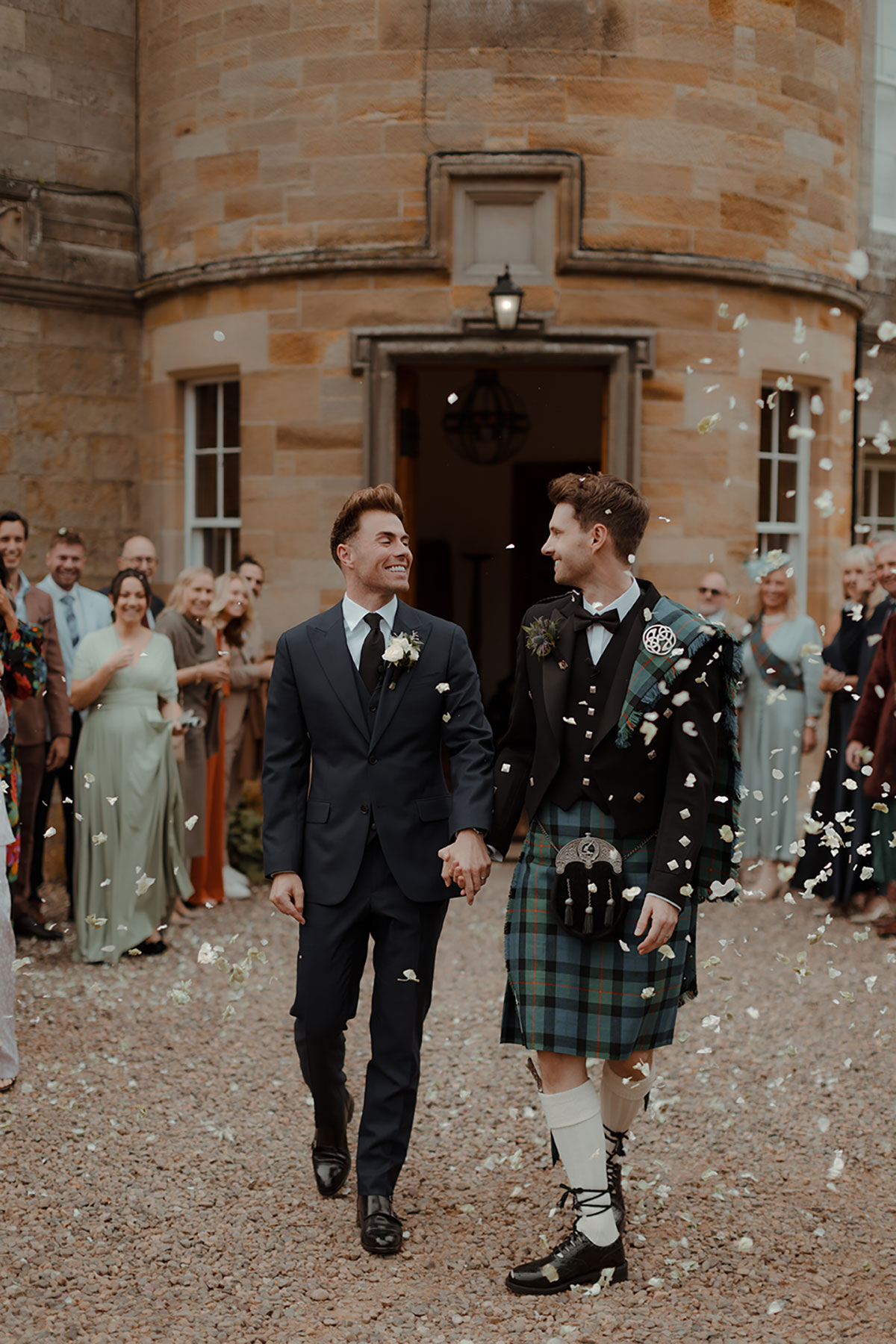 Two grooms walk hand in hand through falling confetti outside Newhall Estate, wearing a wedding suit and a Gunn tartan kilt by MacGregor and MacDuff