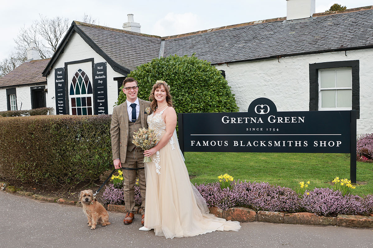 bride and groom stand next to gretna green welcome sign while holding border terrier on lead
