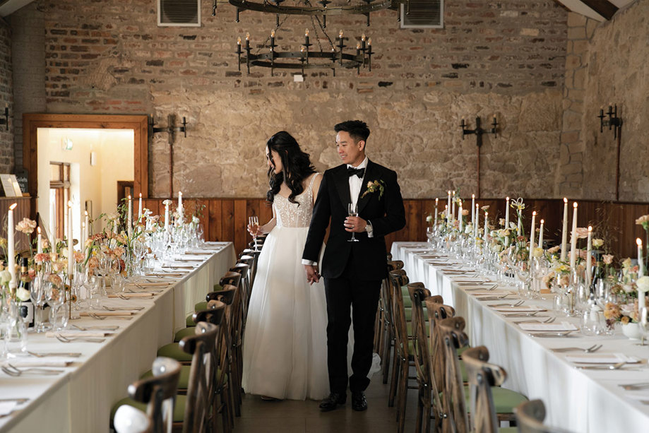 A bride and groom holding hands walking past long tables in a stone barn