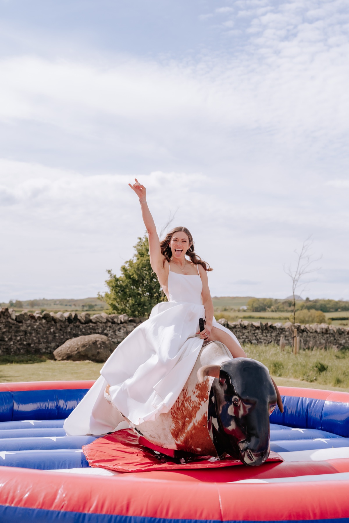 Bride riding a mechanical bull in her wedding dress during Hannah and Donna’s Harvest Moon wedding.