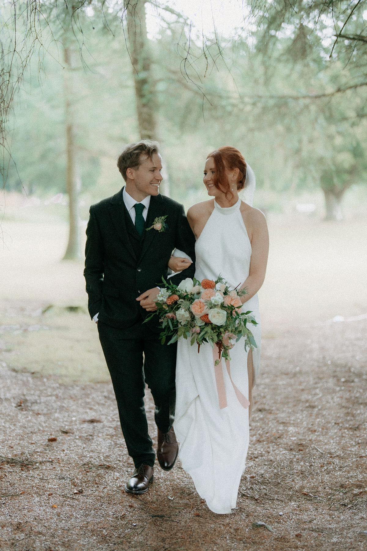 Bride and groom walking arm in arm through woodland holding peach and white wedding bouquet during outdoor Scottish weddin