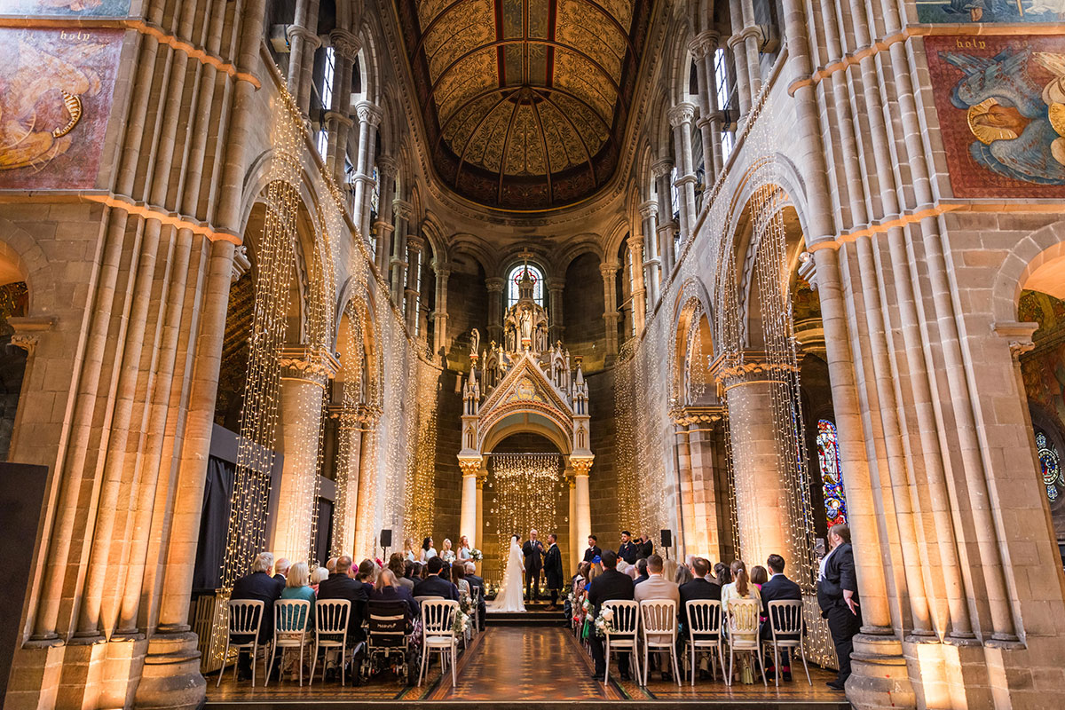 Wedding ceremony taking place beneath the ornate arches and murals of Mansfield Traquair, decorated with strings of warm fairy lights.