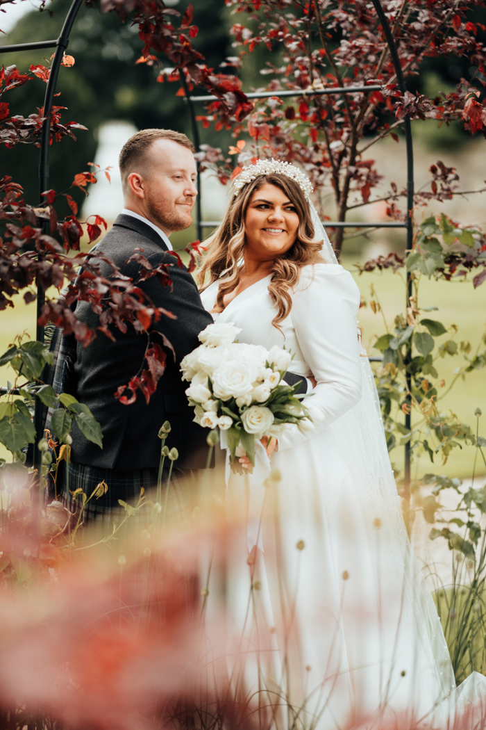 A bride and groom stand together posing for a photo in a garden under an archway 