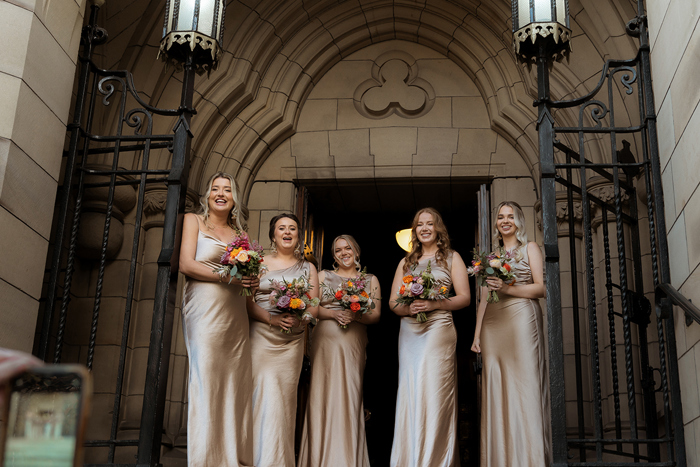 Five Bridesmaids Standing Outside A Stone Building With Black Iron Gates Wearing Oyster Satin Bridesmaid Dresses