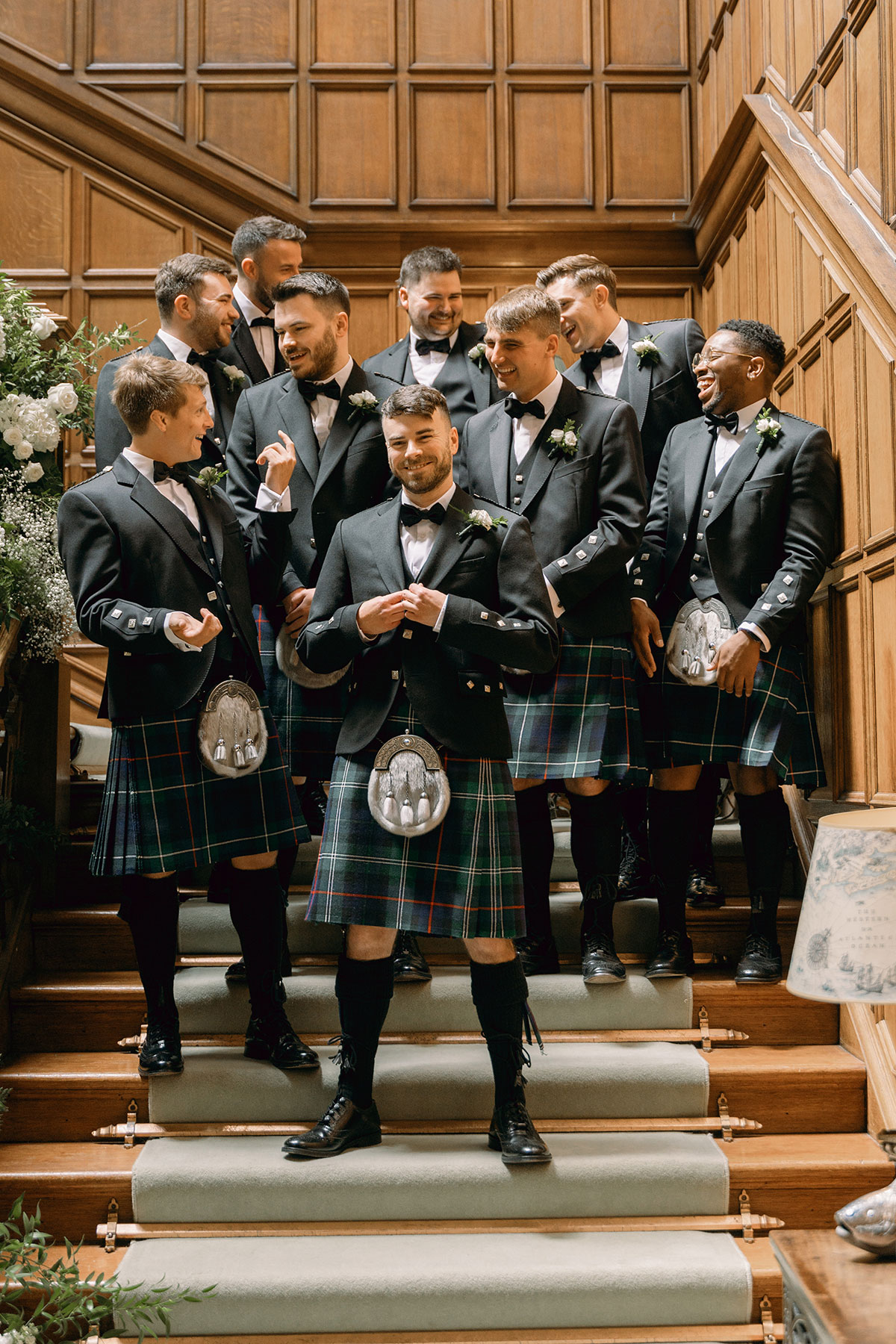 Groom and groomsmen in kilts laugh together on wood-panelled staircase inside Dundas Castle.