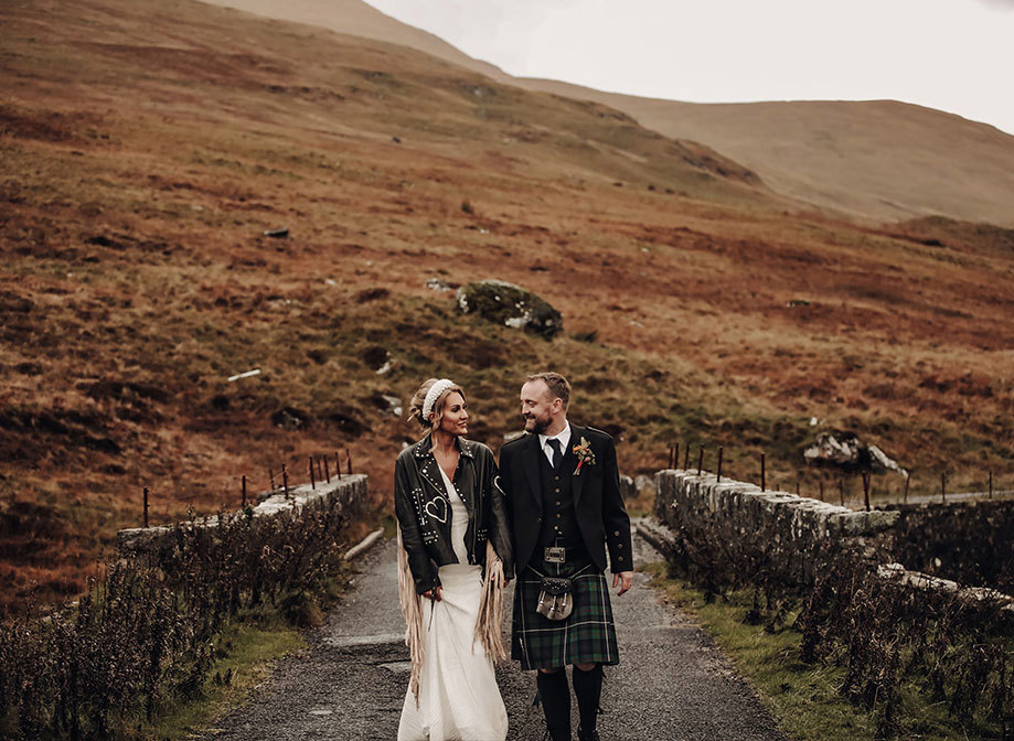 a bride wearing a fringed leather jacket over her wedding dress walking with a groom wearing a kilt along a stone path amid autumn hills and countryside