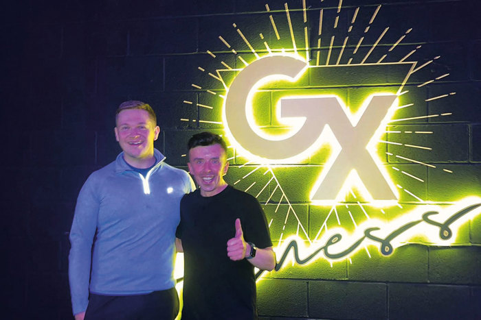 Two men stand in front of a neon yellow sign that says 'GX Fitness' while wearing gym clothes