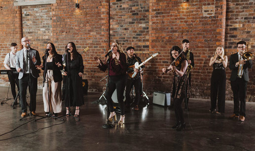 a large group of musicians each playing a different instrument together stand in a large unfurnished room with a large brick wall behind them