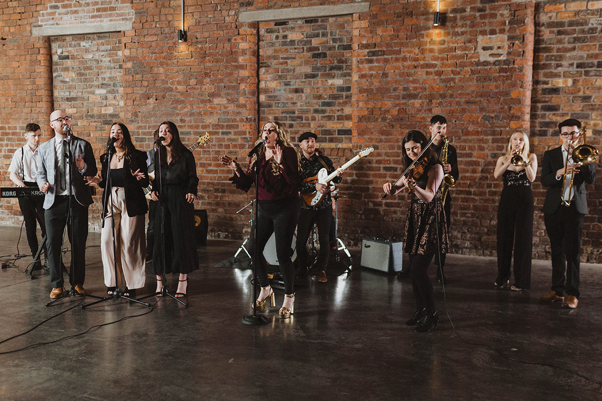 a large group of musicians each playing a different instrument together stand in a large unfurnished room with a large brick wall behind them