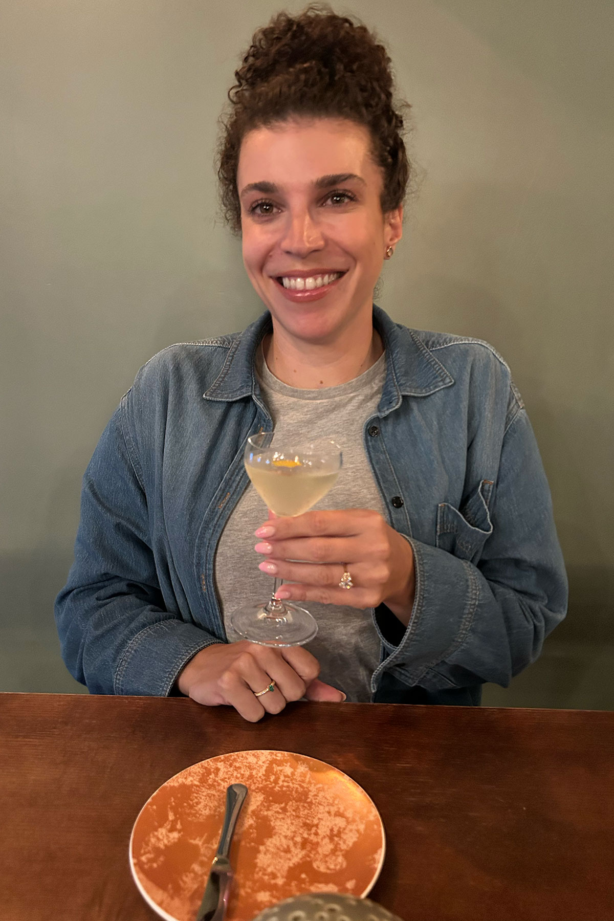 Woman smiling while holding a cocktail at a restaurant table, wearing a denim jacket and engagement ring