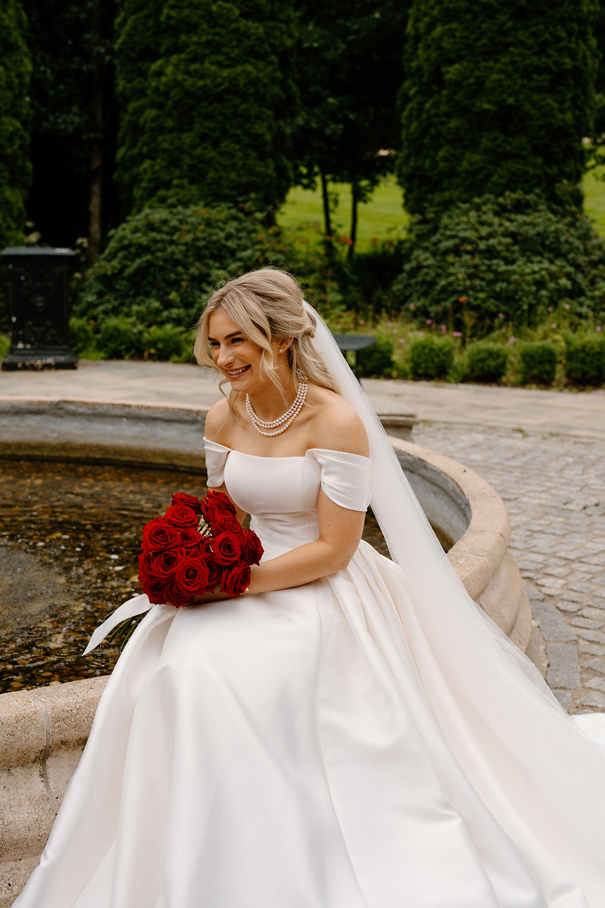 Bride laughing beside fountain holding red rose bridal bouquet during outdoor wedding portraits