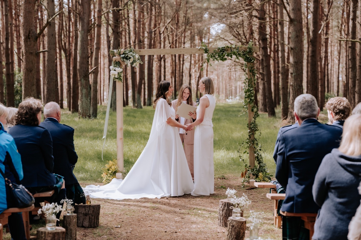 Hannah and Donna marrying beneath a greenery-decorated arch during their woodland ceremony at Harvest Moon.