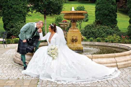 a groom kissing a bride's hand. They are posing by a fountain in a garden.