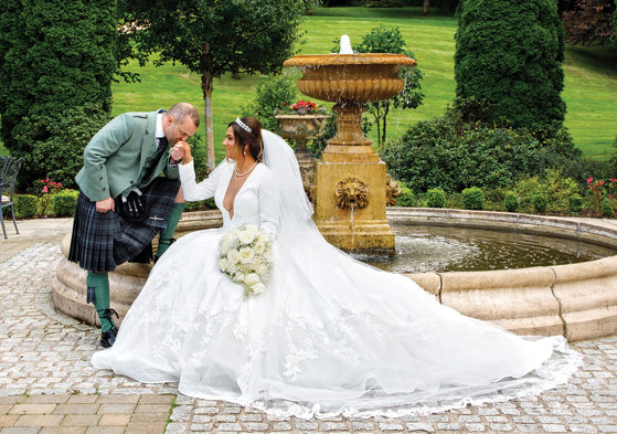 a groom kissing a bride's hand. They are posing by a fountain in a garden.