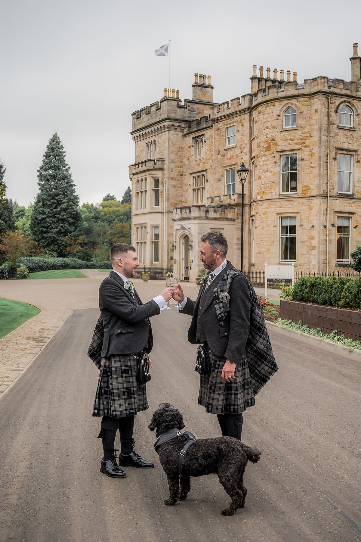 Same-sex couple in kilts toasting outside Crossbasket Castle with their dog on their wedding day