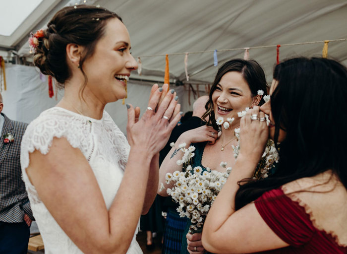 an elated bride wearing a fine lace top clasps her hands while talking to two other wedding guests in a marquee. They are holding bouquets of white daisies and there are colourful paper ribbon streamers hanging from a rope in the background 