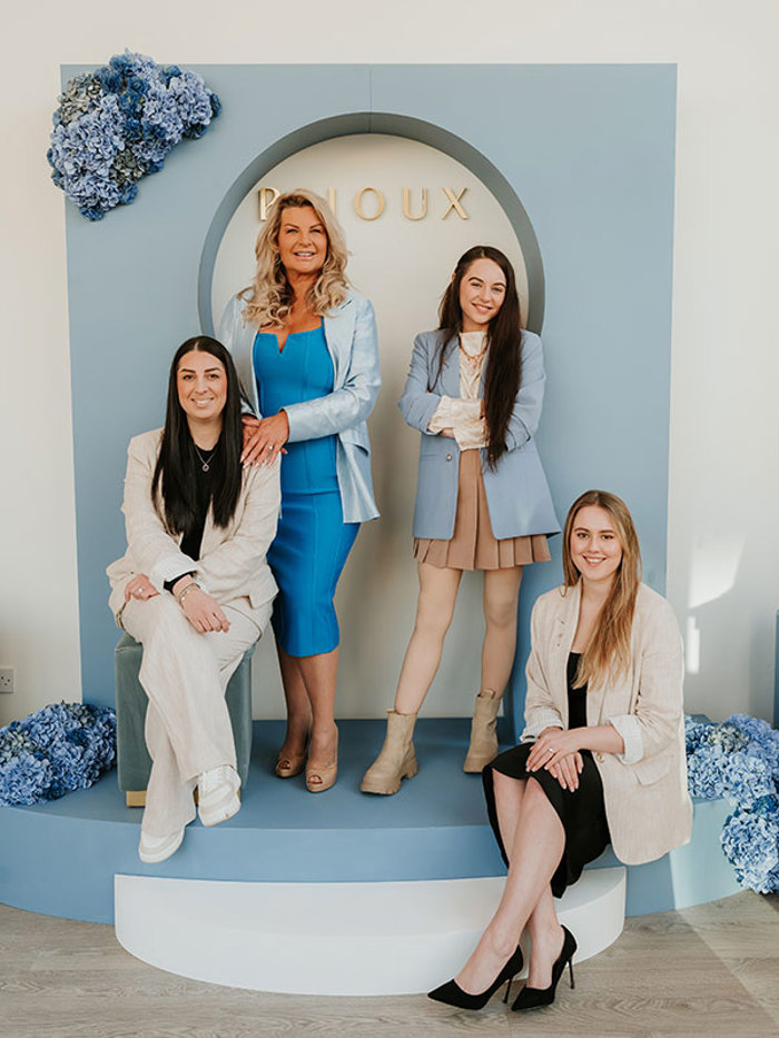 four women sat down and standing on a blue platform and background that says Bijoux in gold letters in the back