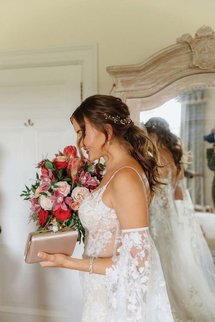 side view of bride in floaty floral embroiderd corset bodice wedding dress stood in front of a red and pink flower display and a large wardrobe mirror
