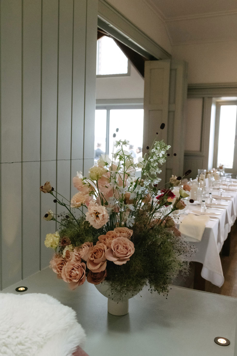 a large pastel flower arrangement sitting on a grey surface with dinner table in background