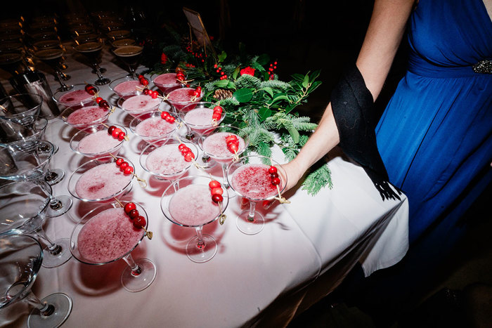 rows of red cocktails in martini glasses topped with a three skewered cranberries each are placed on a white table cloth next to a pine tree decoration