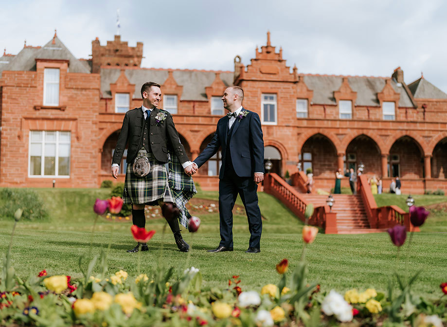 two grooms holding hands looking at one another walking through a garden with red sandstone exterior of Boclair House in the background