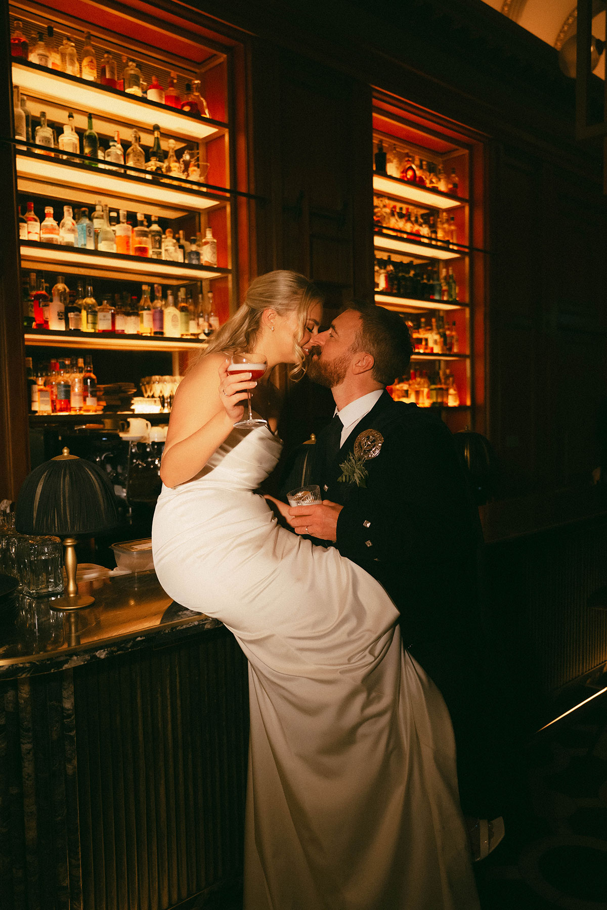 bride sitting on bar kissing groom with cocktails in stylish wedding reception bar