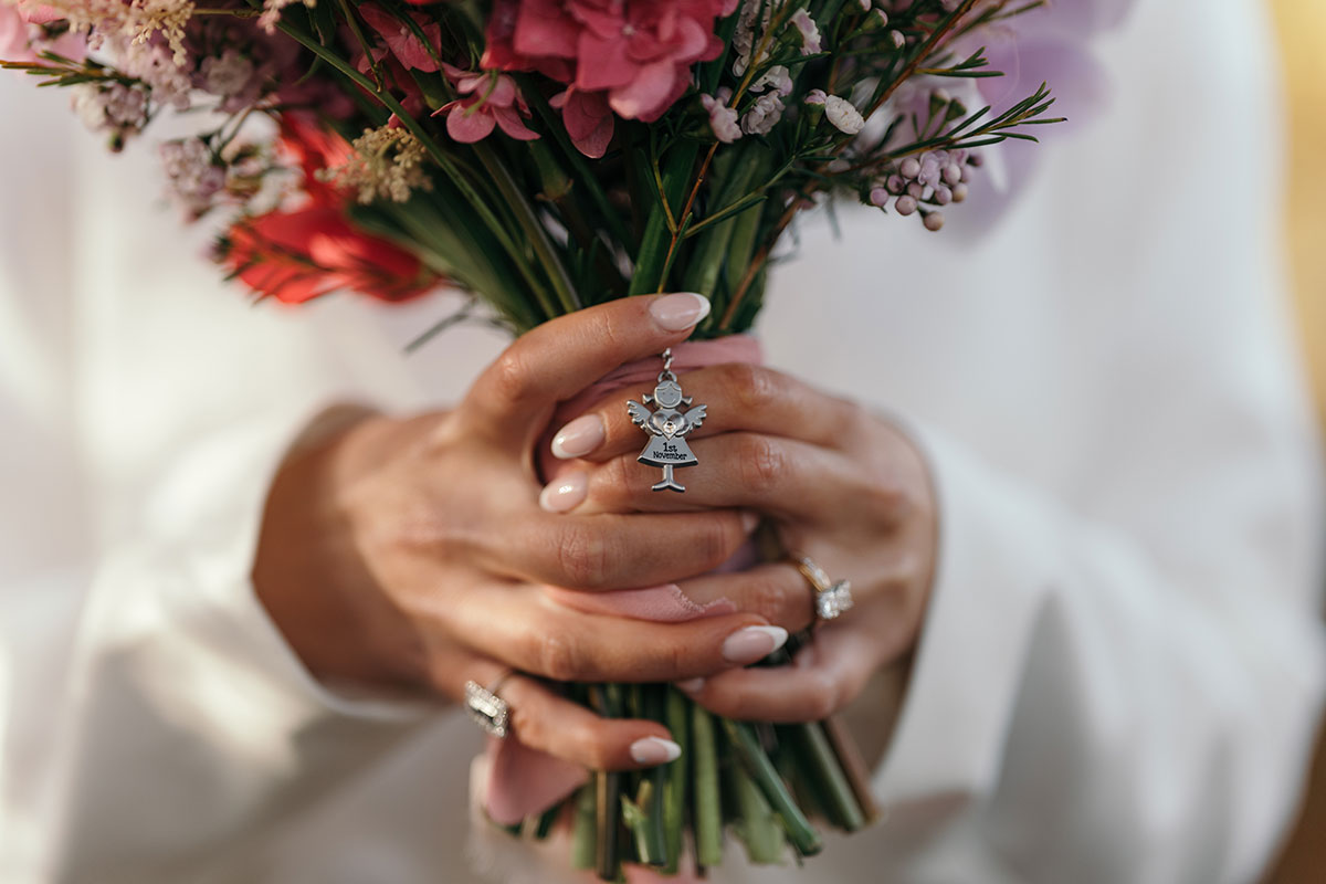 Close-up of bride’s hands holding pink wedding bouquet with rings and personalised charm detail