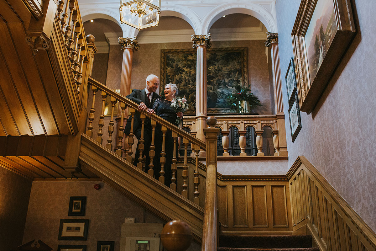 Couple smiling together on the grand wooden staircase at Norton House Hotel, Edinburgh