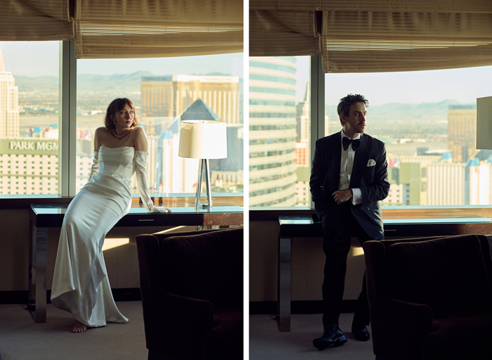 A woman in a white gown and a man in a tuxedo pose in a hotel room, overlooking a vibrant cityscape through large windows.