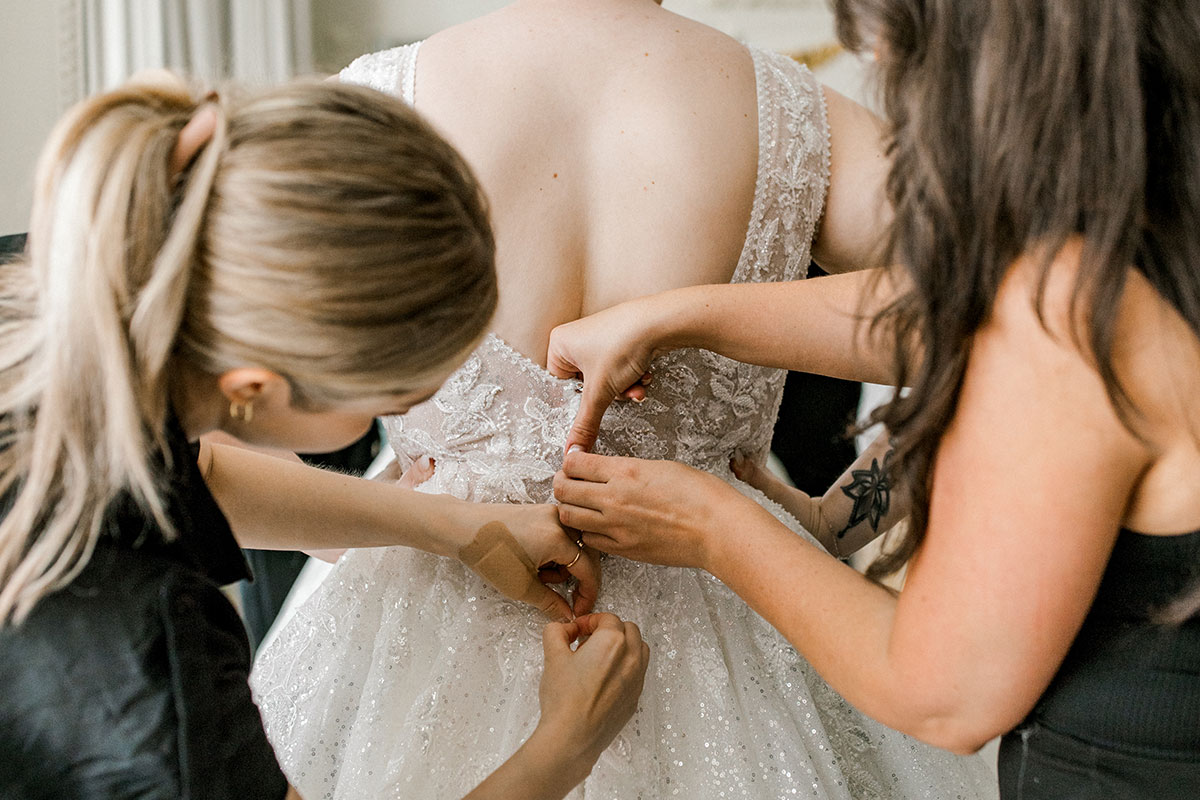 blonde and brunette women help button up the back of bride's wedding dress, one of them stood on either side