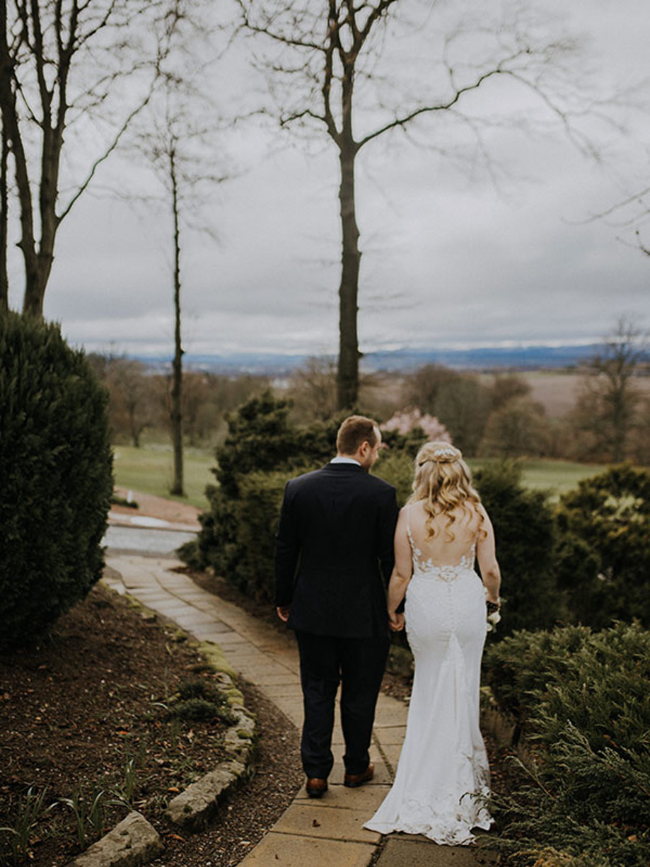 A man in a suit and a woman in a wedding dress walk along a path away from the camera 