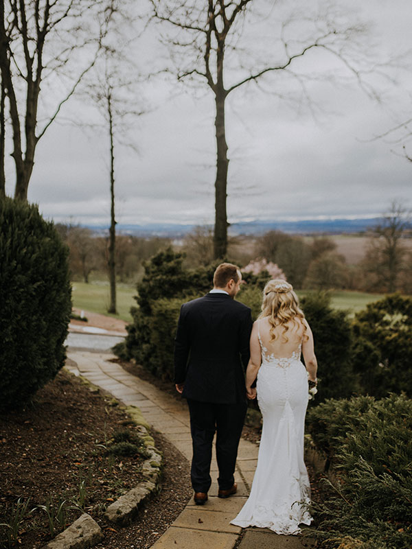 A man in a suit and a woman in a wedding dress walk along a path away from the camera