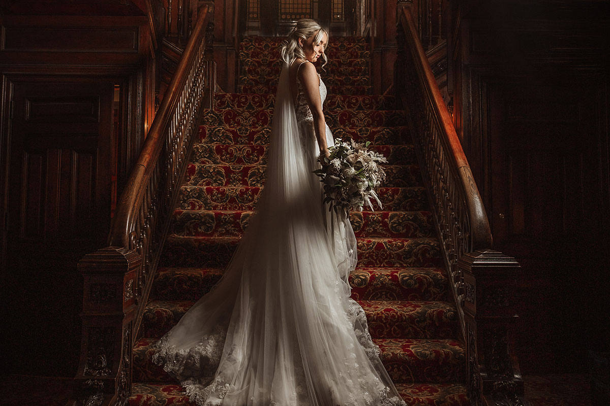 Bride in lace gown holding bouquet on ornate staircase with red patterned carpet and wooden bannisters
