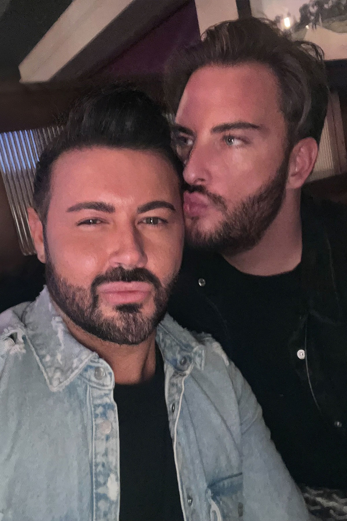 couple close-up selfie with one partner kissing the other on the cheek in a relaxed indoor setting