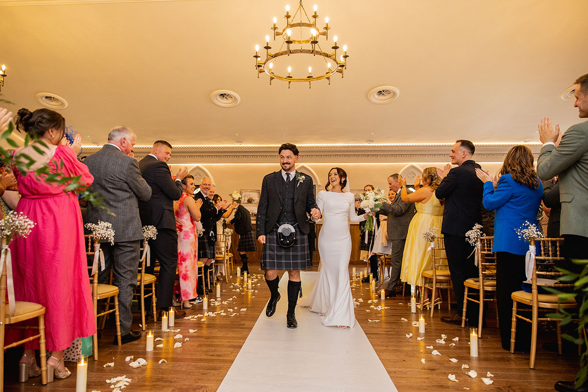 Bride and groom walk up the aisle hand-in-hand as guests cheer and applaud their ceremony