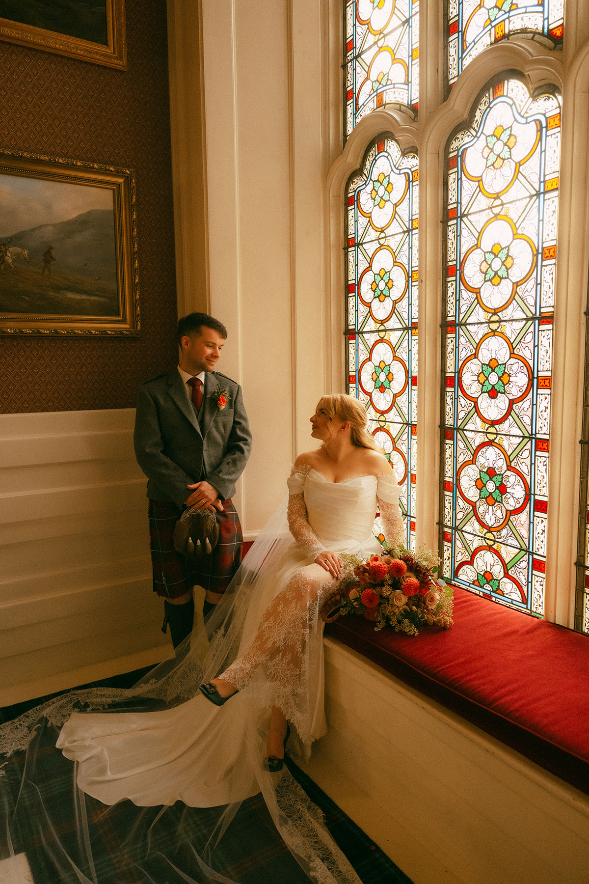 Bride seated by stained glass window with bouquet while groom stands nearby inside elegant Scottish wedding venue