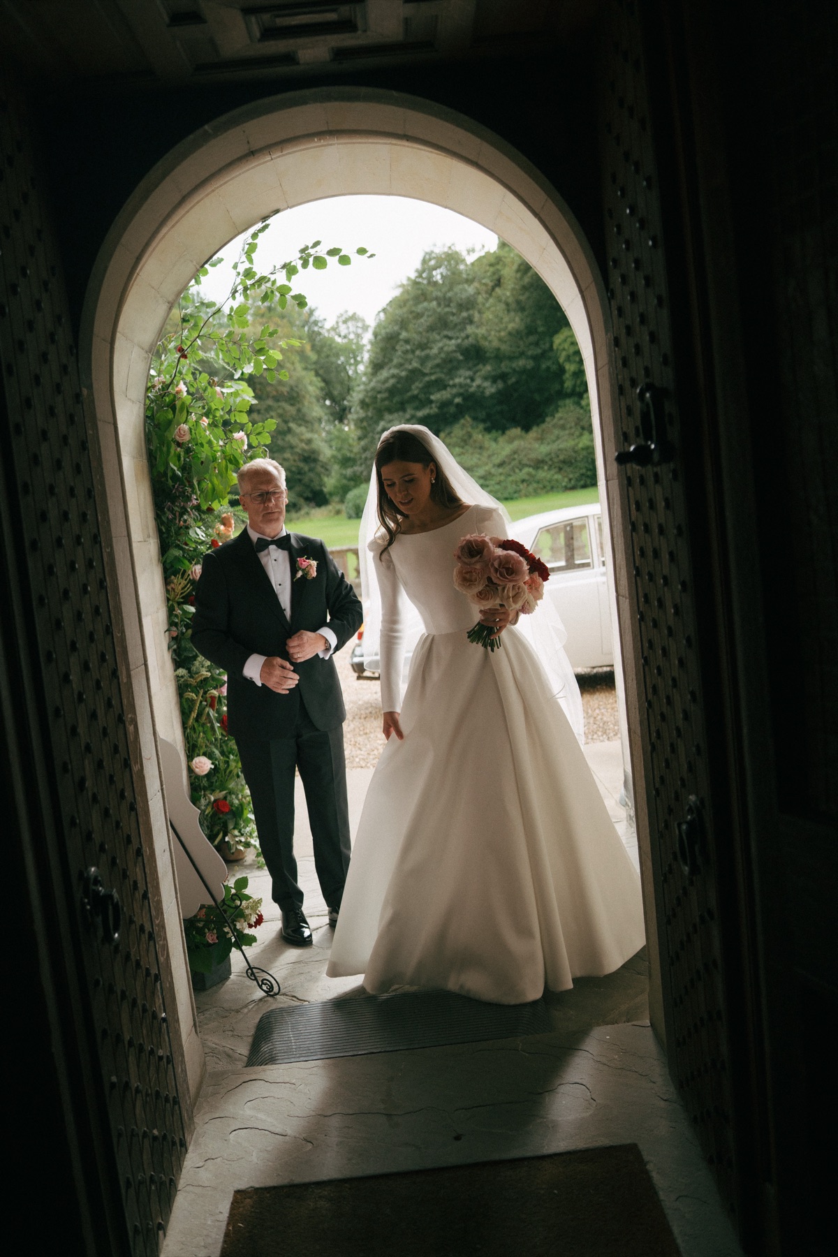 Bride Nicole enters Barnbougle Castle with her dad, wearing a bespoke Suzanne Neville wedding dress and carrying a pink-and-red bouquet.