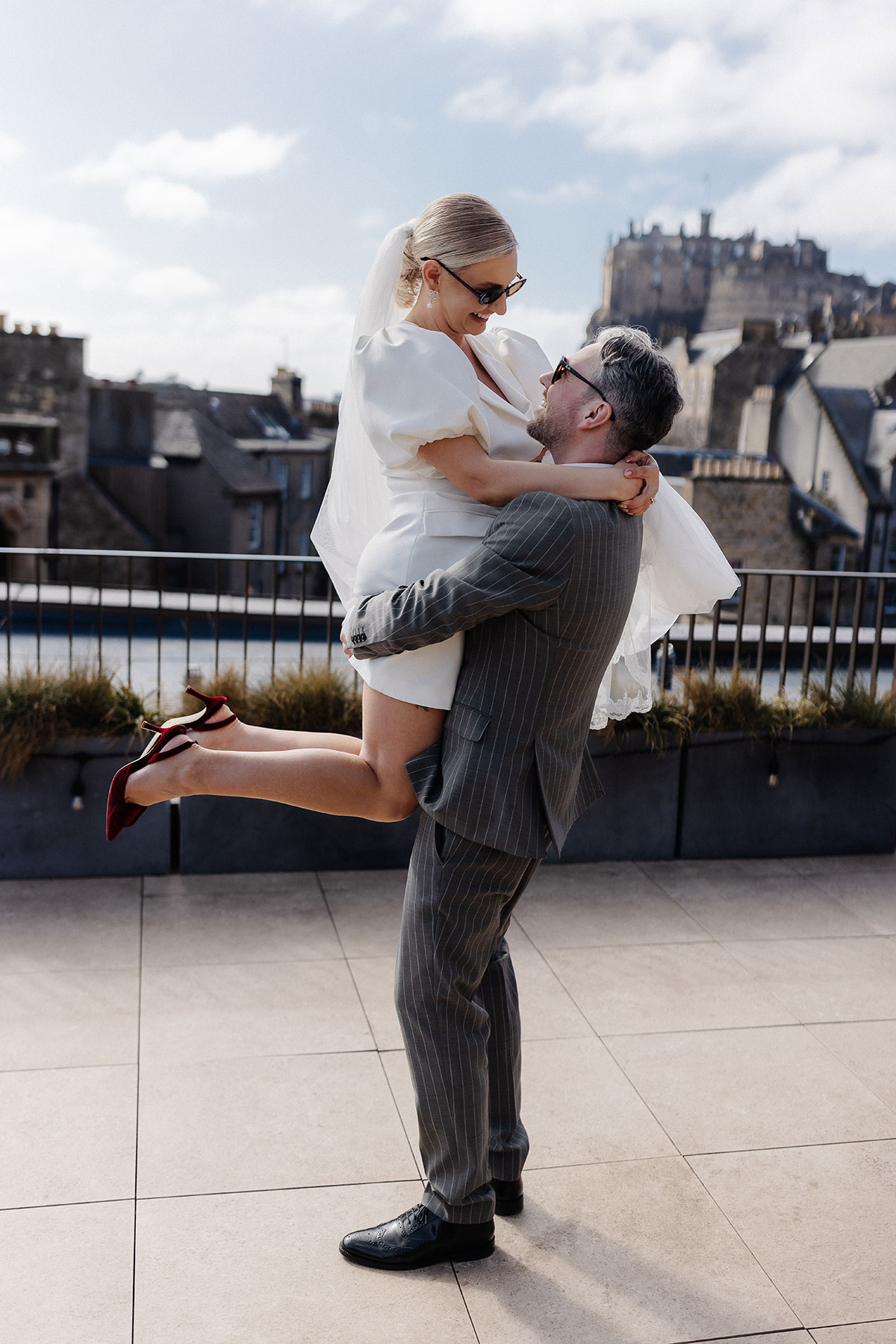 Groom lifts bride in a playful moment on the Virgin Hotels Edinburgh terrace, the castle skyline behind them on their modern city wedding day.